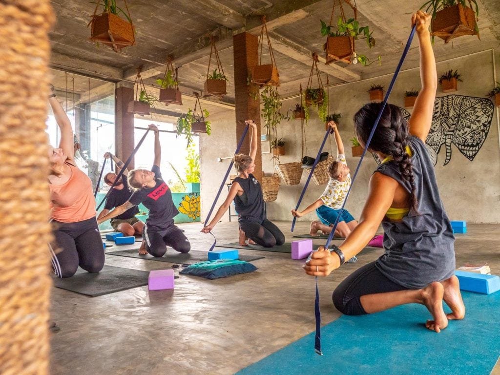 A group of people engage in a yoga class using straps suspended from the ceiling, with hanging plants and a mural in a spacious, airy room. Yoga is a must-do thing in Ahangama
