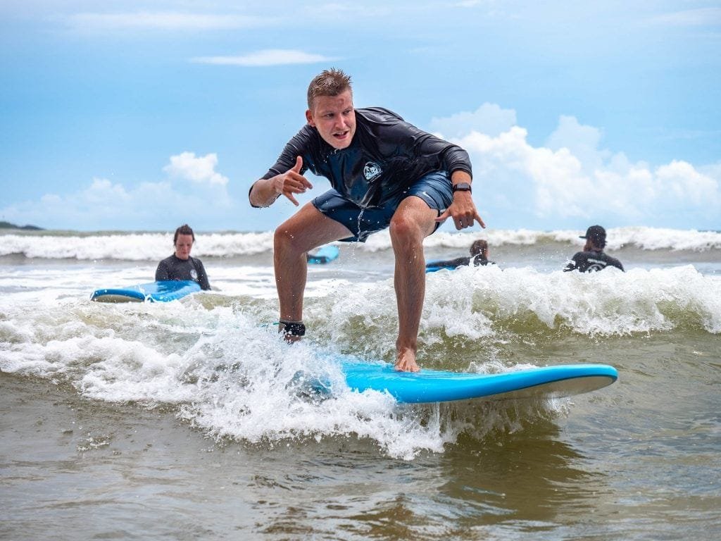 A man in a black wetsuit is surfing a blue wave, intensely focused as he crouches on his board, with other surfers visible in the sea behind him.