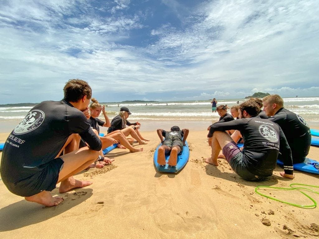 A group of people in black rash guards are sitting on surfboards on the beach, attentively watching a surf instructor demonstrate a technique.