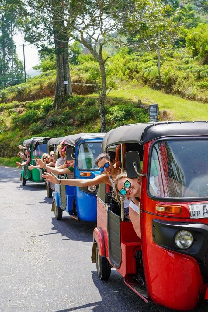 Tourists in tuk-tuks waving and smiling on a sunny road lined with trees, showcasing a fun and scenic travel experience.