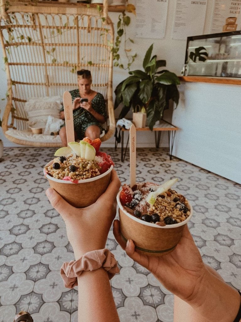 Two hands holding bowls of fruit and granola bowls inside a cozy café with a hanging chair and potted plants in the background.