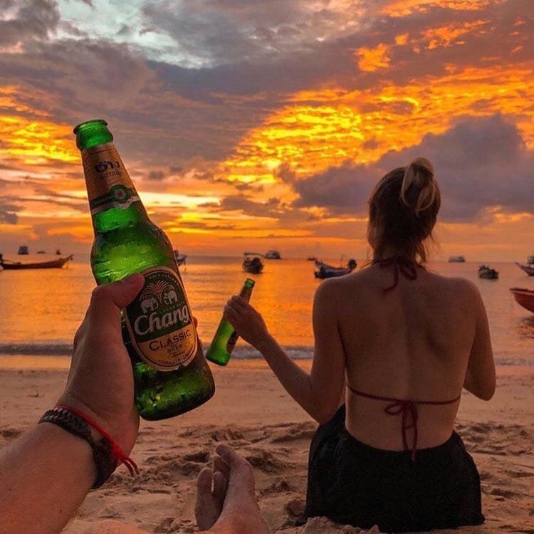 A couple sat on the beach in Thailand with beers in hand enjoying the sunset