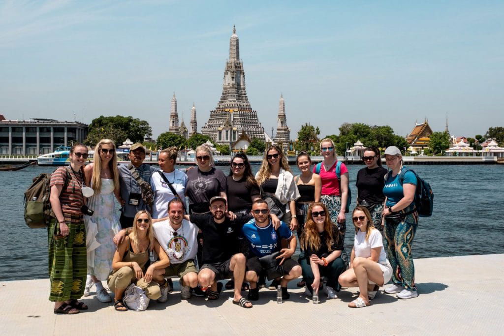 A large group of tourists posing in front of the iconic Wat Arun temple across the river under a clear blue sky. 
