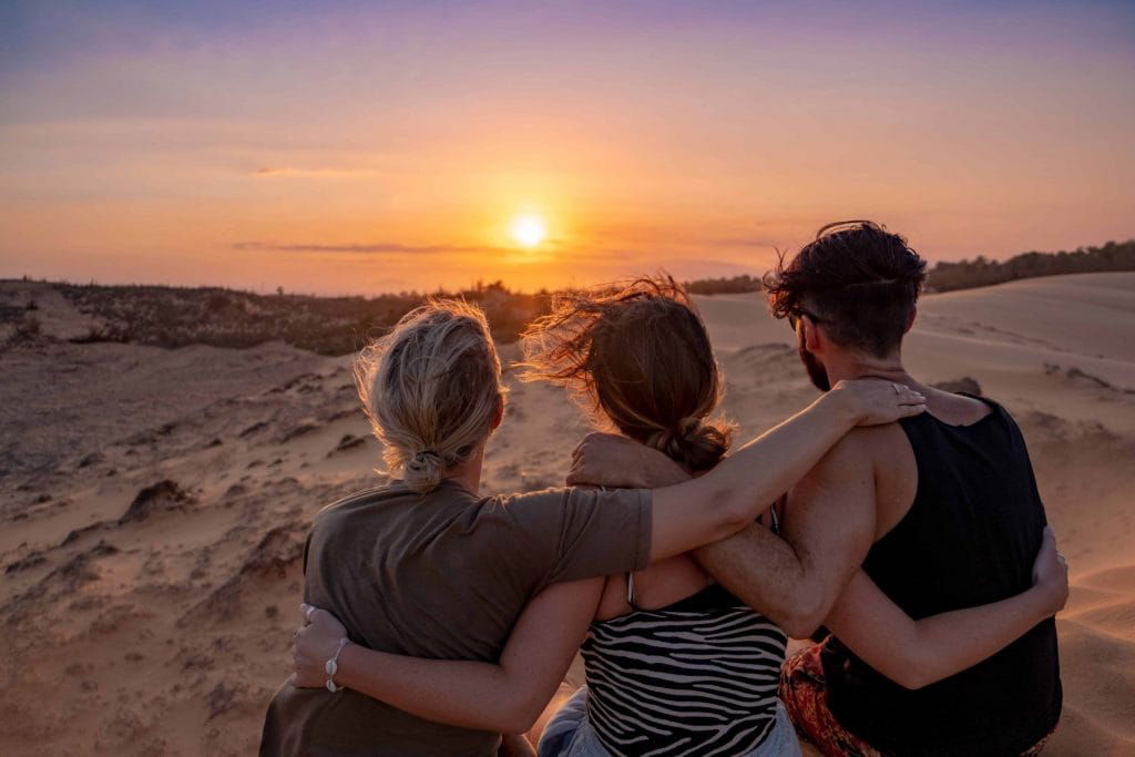 Three friends watching a stunning sunset, embracing each other on a sandy desert with vast dunes under a vibrant sky.
