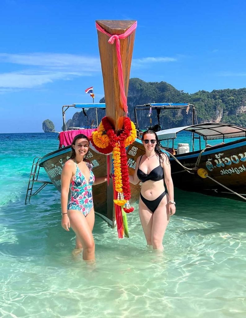 Two girls standing by a long boat in the water while island hopping.