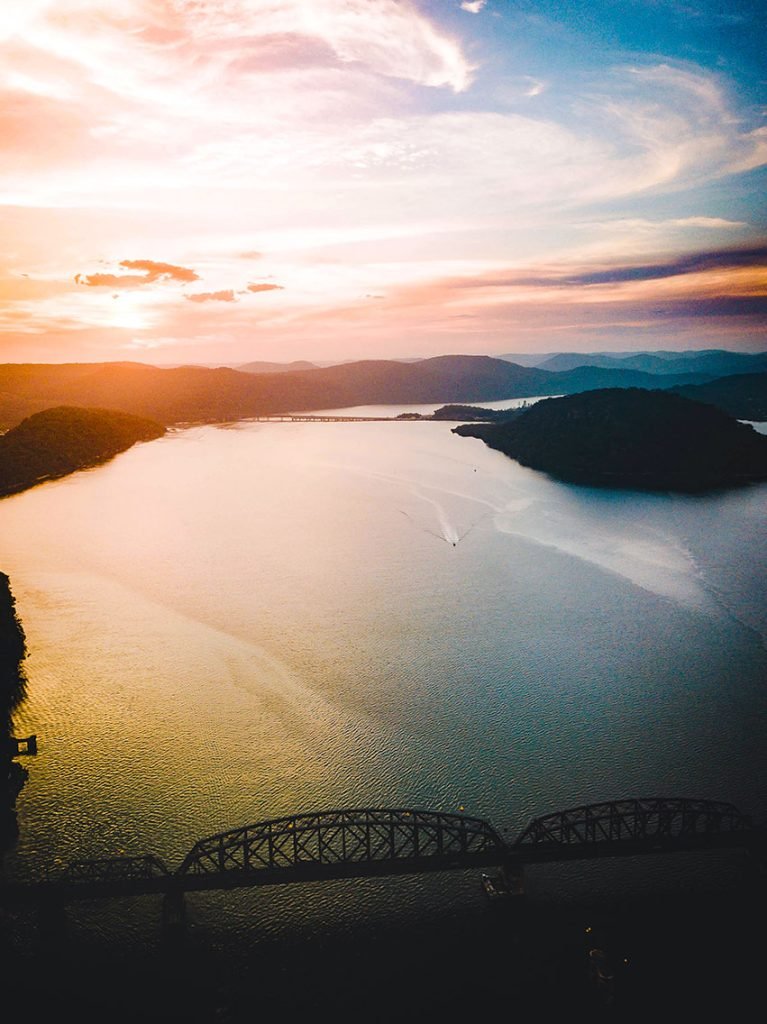 Sunset hues paint the serene Hawkesbury River, bordered by the Australian bush and a railway bridge silhouette in the distance. It's one of the best day trips from Sydney.