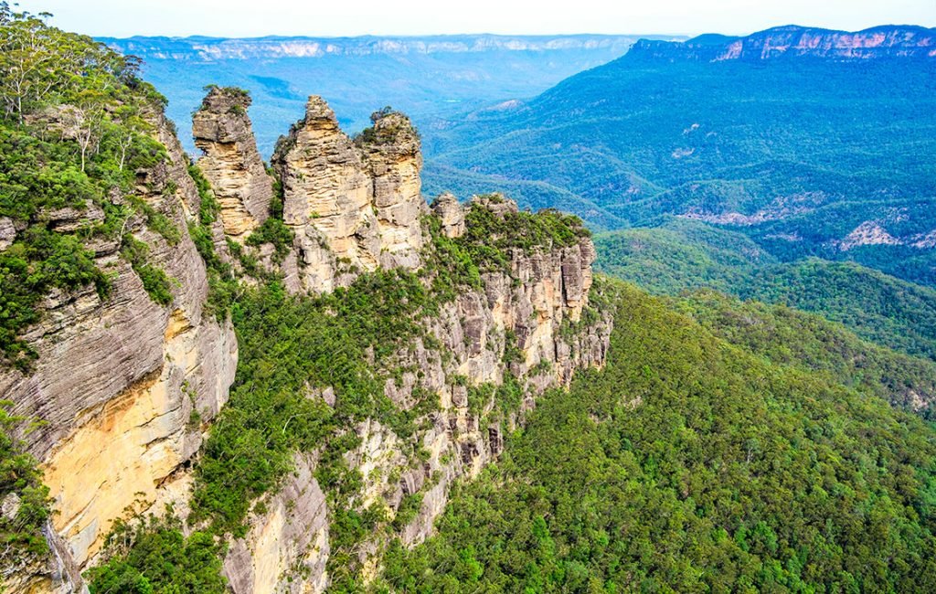 The iconic Three Sisters rock formation towering over the verdant Jamison Valley in the Blue Mountains, Australia, under a clear sky.