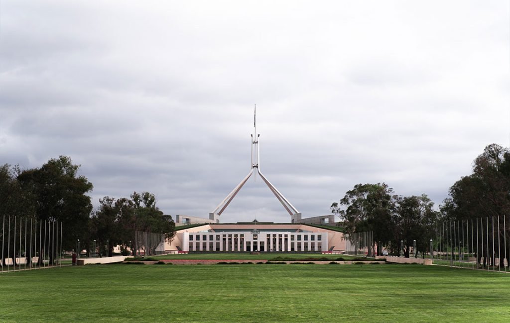 The modern architecture of Parliament House in Canberra, Australia, seen from the lush front lawns, under an overcast sky.