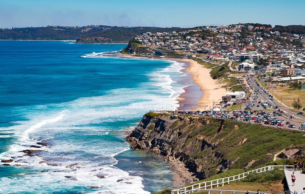 Vibrant scene of Newcastle Beach with surfers in the water, backed by a bustling cityscape and coastal road in New South Wales, Australia.