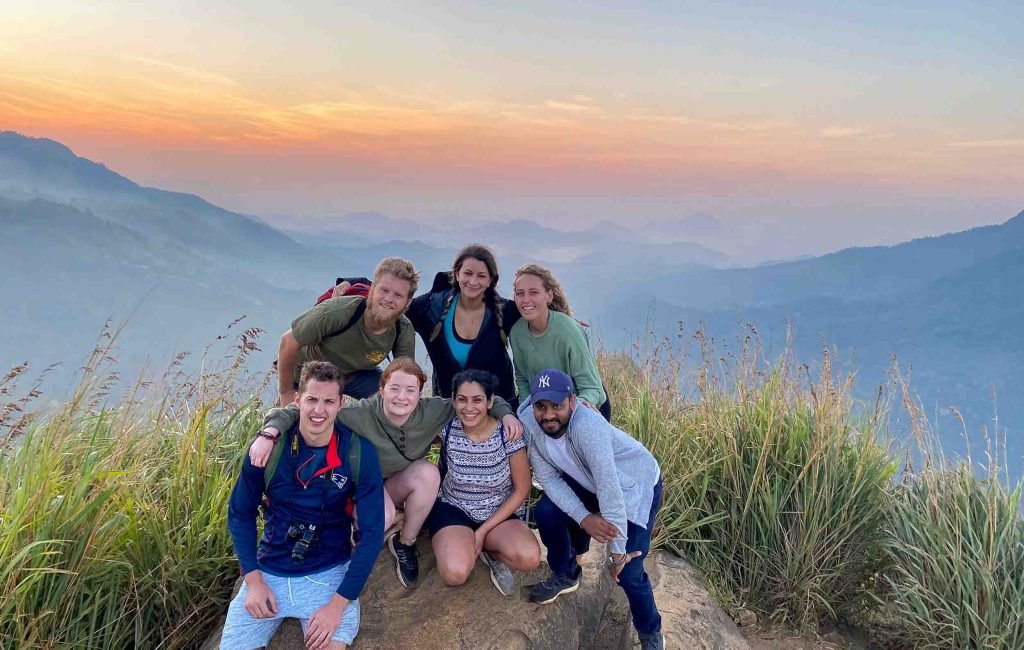 A group of smiling hikers at the summit during sunrise with expansive mountain views in the background