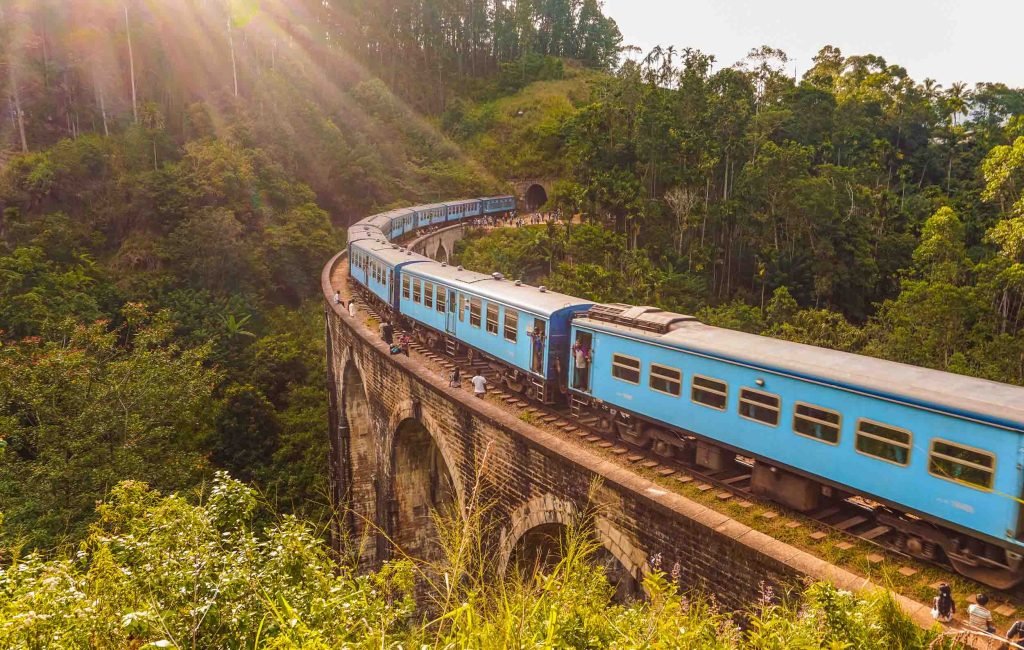 A train passing over the Nine Arches Bridge in Sri Lanka, surrounded by lush greenery and bathed in sunlight, with the hills in the background.
