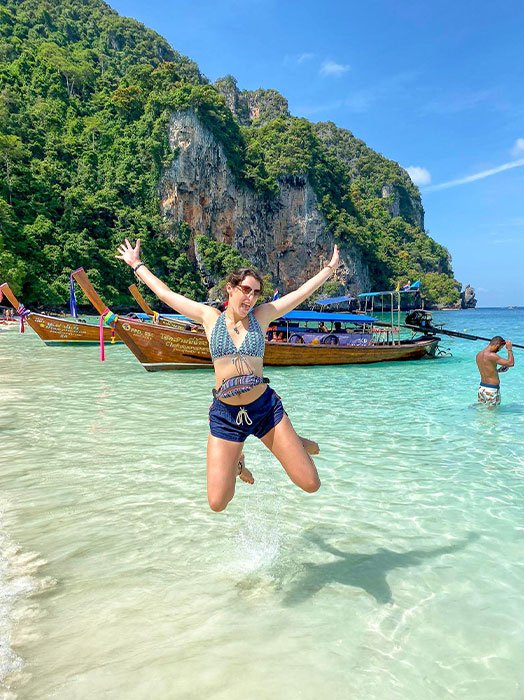 A happy solo traveller in summer wear jumping on a pristine beach in Krabi with clear waters, traditional longtail boats, and a rocky cliff in the background, conveying the excitement of a tropical solo backpacking holidays.