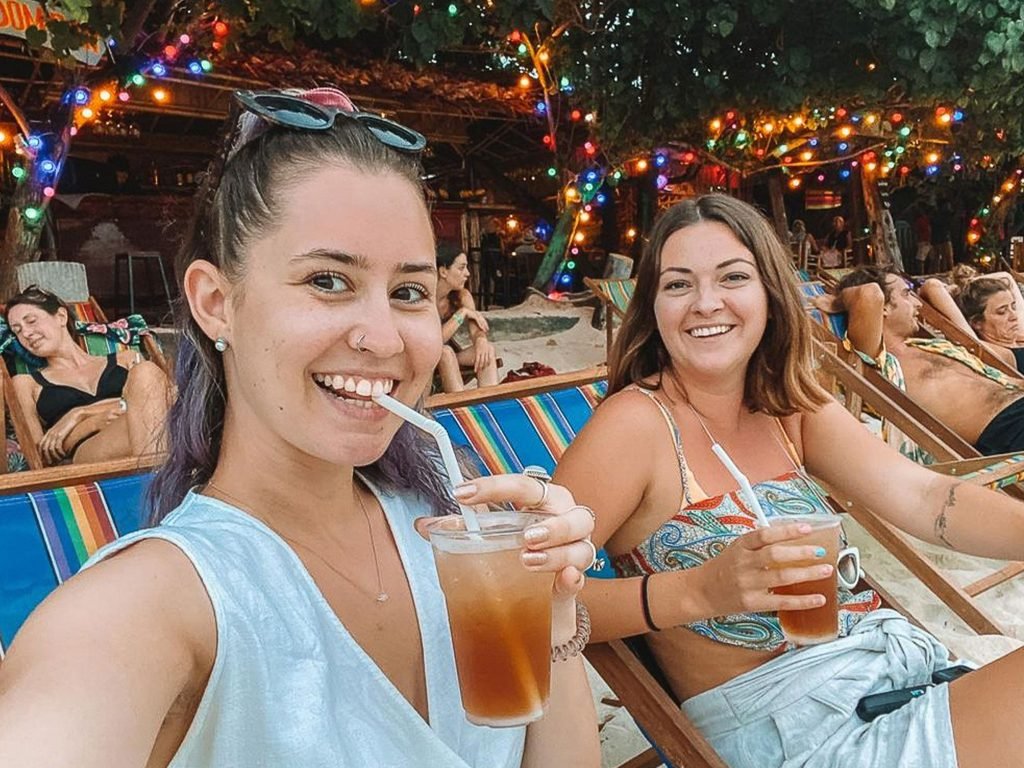 Two solo travellers smiling for a selfie while relaxing on colourful beach chairs, enjoying iced drinks, with string lights and fellow travellers in the background
