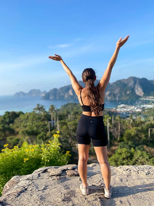 A solo traveller stands with her back to the camera, arms raised overlooking a panoramic view of a the Phi Phi islands with limestone karsts in the distance.