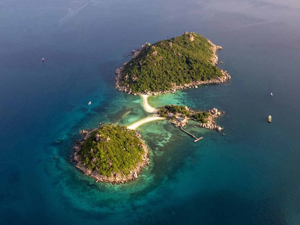 Aerial perspective of a tropical island near Koh Tao with two lush green islands connected by a sand bar, surrounded by turquoise waters and boats near the shore.