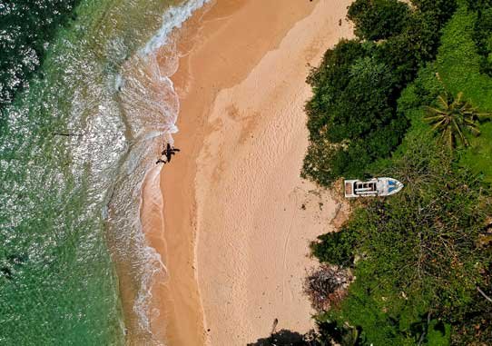 Aerial view of a pristine beach with a lone boat ashore and the waves gently breaking at the edge of the lush greenery