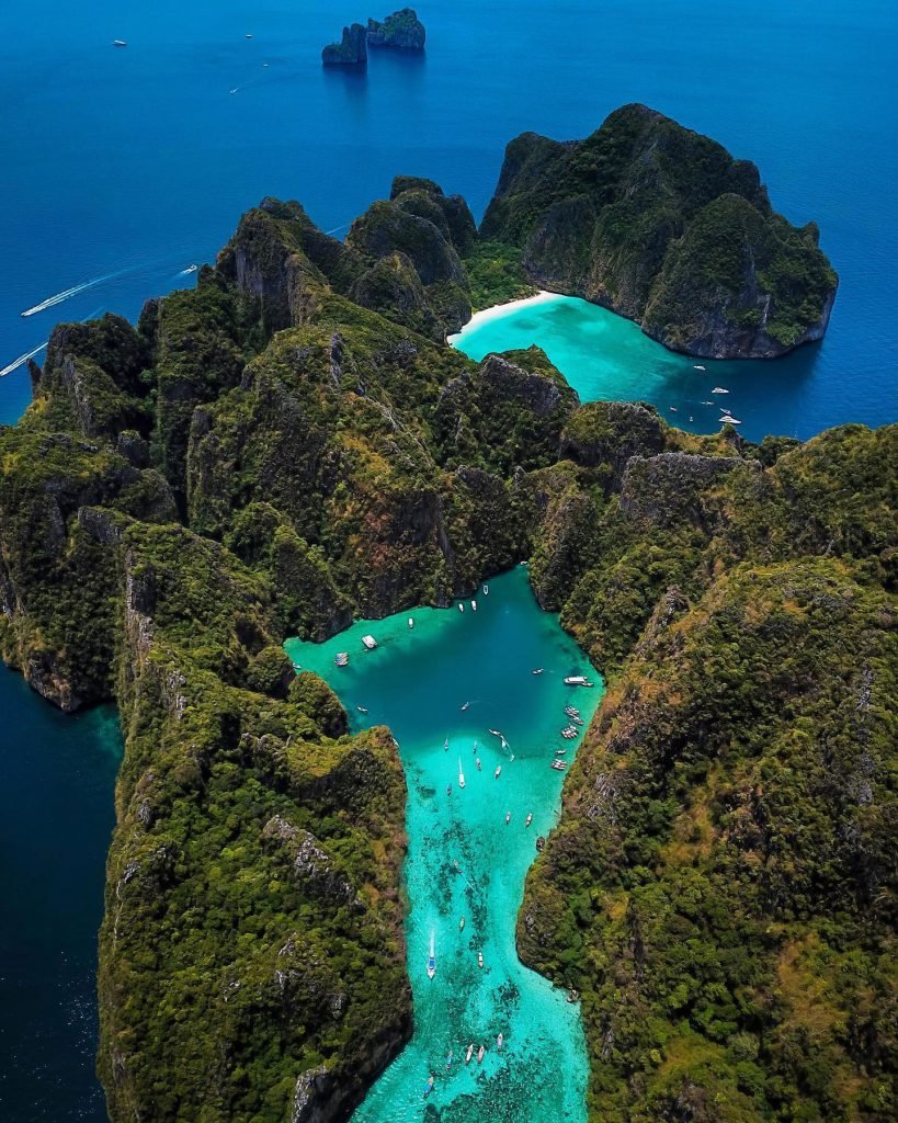 Aerial view of a hidden lagoon in Phi Phi with turquoise waters nestled between emerald-green karst cliffs, with boats scattered around, showcasing the beauty of a tropical paradise.