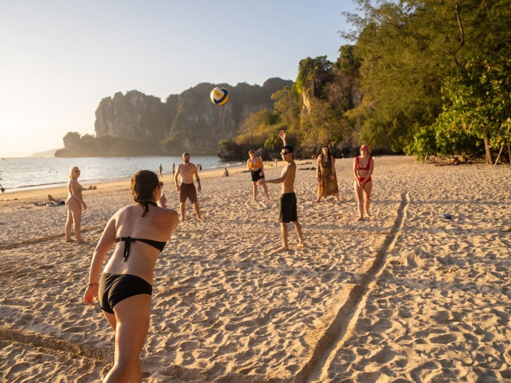 Group of backpackers enjoying a game of beach volleyball at sunset, with the ball mid-air, surrounded by a scenic landscape of sandy beach and towering cliffs.