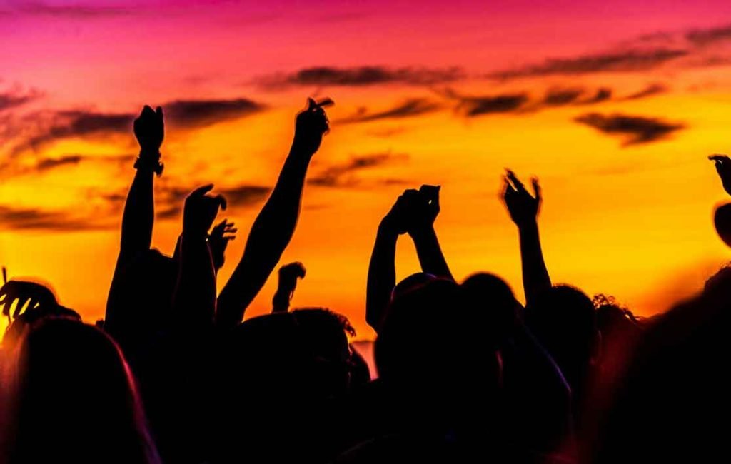 Silhouettes of a crowd of people at a beach party, with arms raised against a vibrant sunset sky. One of the best things to do when island hopping in Thailand is to attend the Full Moon Party.