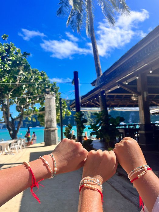 Three individuals' wrists with friendship bracelets against a vibrant backdrop of a tropical beach setting with palm trees and a clear blue sky
