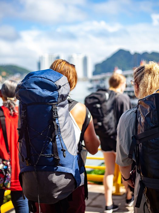 A traveler with a large blue backpack waiting in a queue at a sunny harbour with clear blue skies in the distance