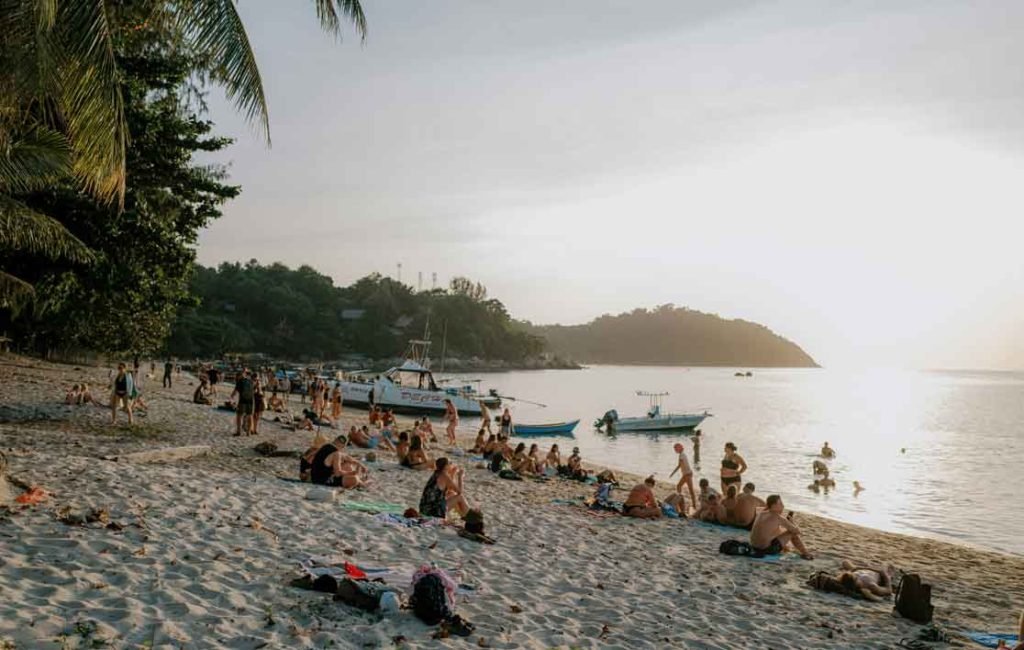 People relaxing on a sandy beach with palm trees and boats in the water as the sun sets, creating a serene evening atmosphere