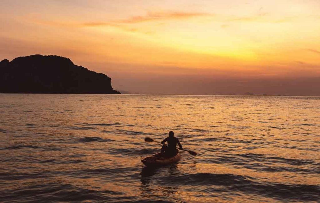Silhouette of a lone kayaker paddling in the ocean at sunset with the silhouette of a mountainous island in the background