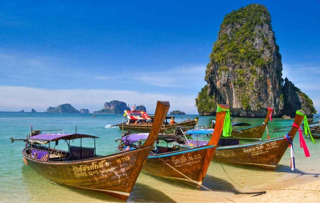 Colorful wooden longtail boats moored on a sandy shore with a limestone karst backdrop under a clear blue sky