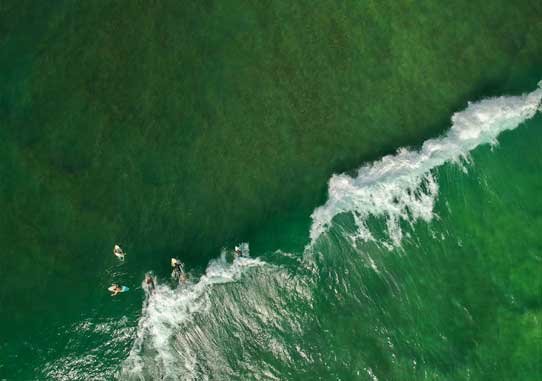 Overhead view of surfers on vibrant green waves, with the surf breaking in a foam, off a sandy beach.