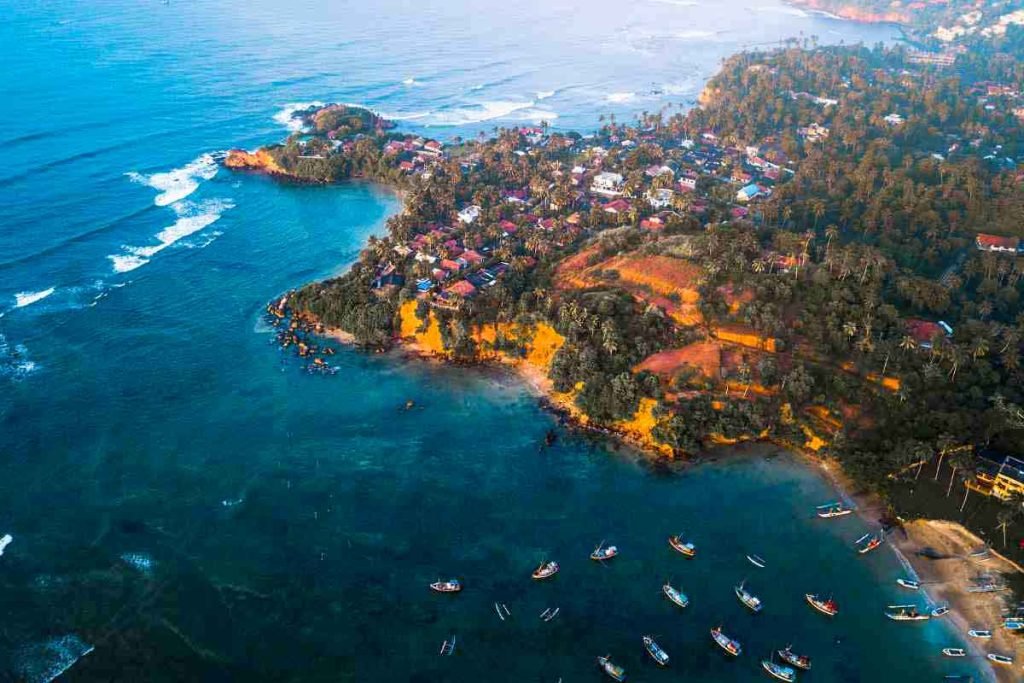 Aerial shot of Weligama with its distinctive coastline, featuring a village nestled among greenery, golden shores, and scattered boats in the bay