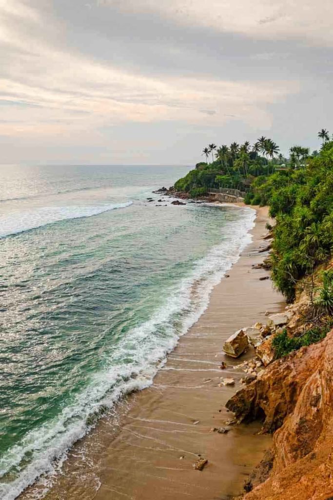 A tranquil view of the Weligama coast with gentle waves washing onto a sandy beach flanked by lush greenery and palm trees