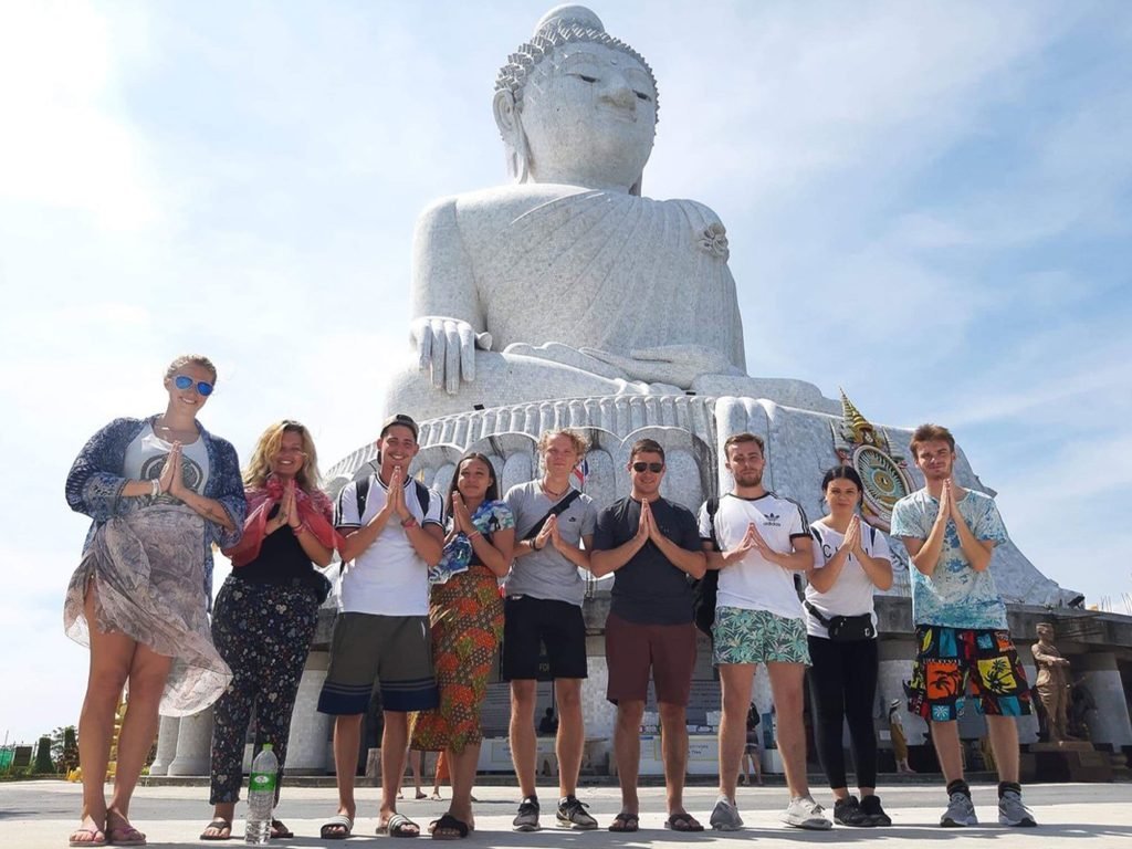 Group of tourists in casual clothing posing with hands clasped in traditional greeting in front of the large, serene Big White Buddha statue under a clear blue sky in Phuket