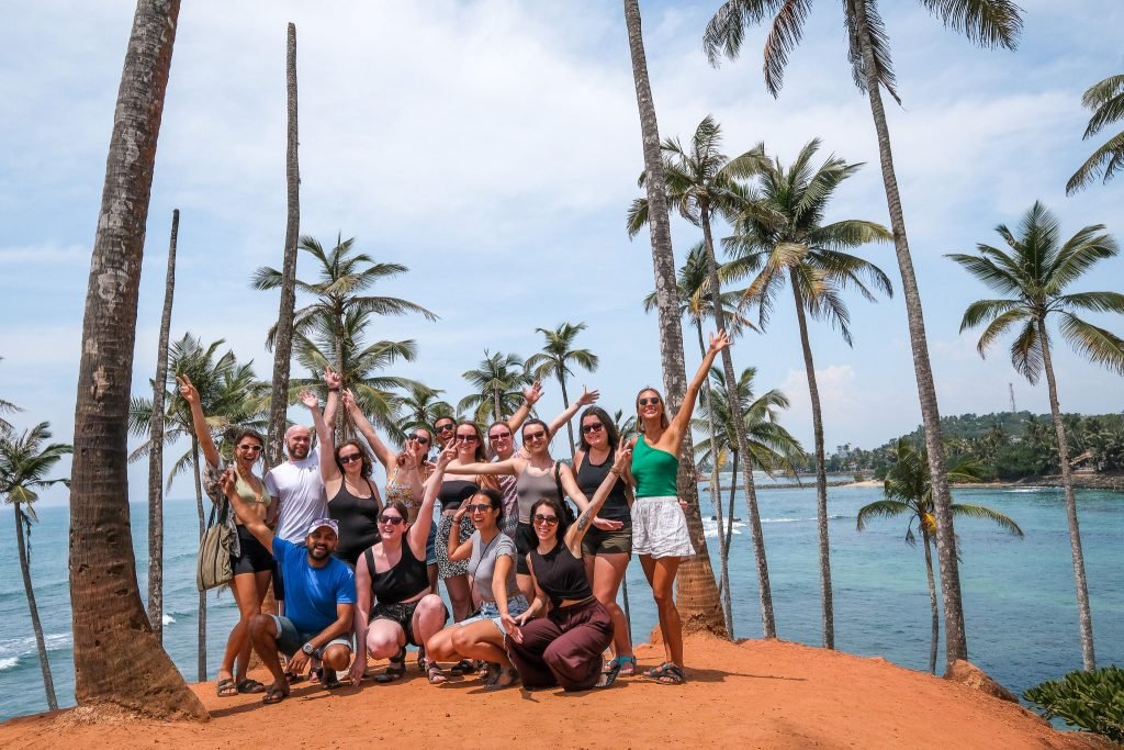 A diverse group of people are smiling and raising their arms in celebration in front of tall palm trees with a tropical beach in the background.
