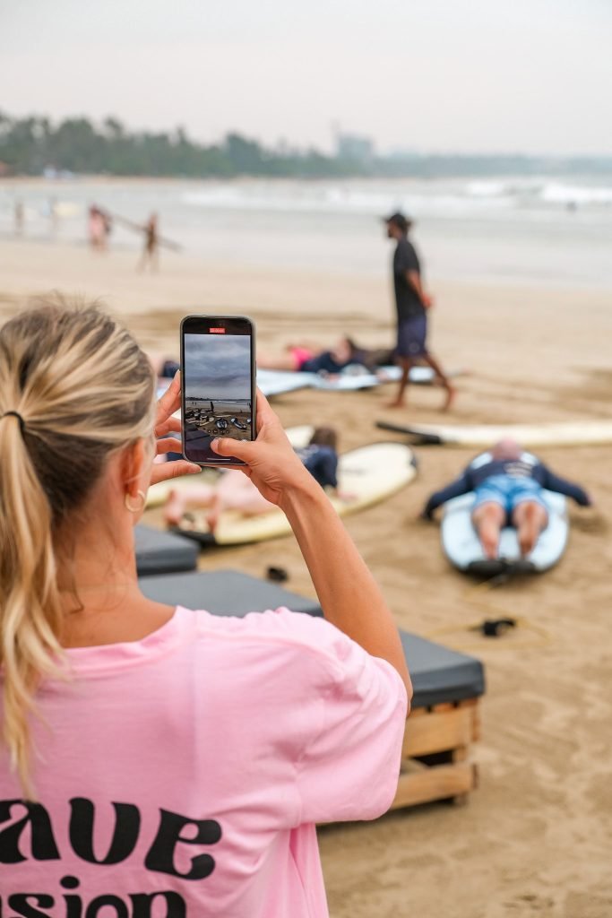 Over-the-shoulder view of a woman using a smartphone to capture a photo of a beach where several people are lying on surfboards. Surfing is one of the best things to do in Ahangama
