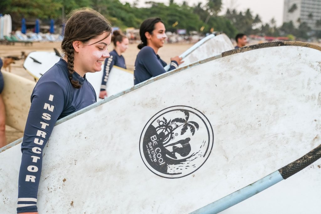 A young solo travellers with a side braid smiles as she carries a white surfboard on a sandy beach, with fellow surfers walking behind her