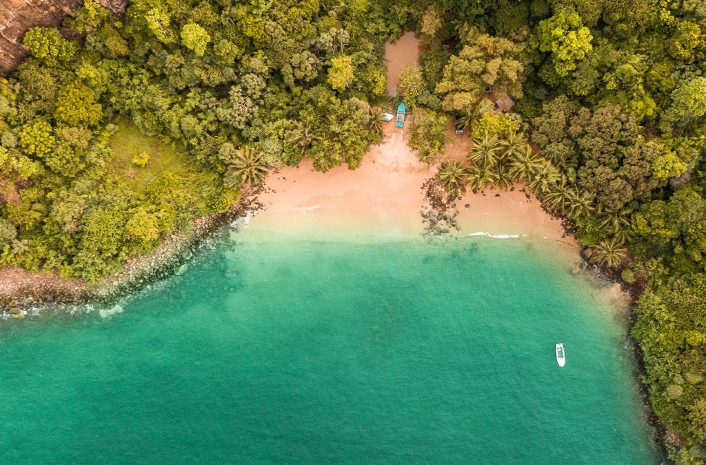 Aerial view of a secluded beach cove with emerald waters surrounded by lush greenery and a single boat moored near the shore.