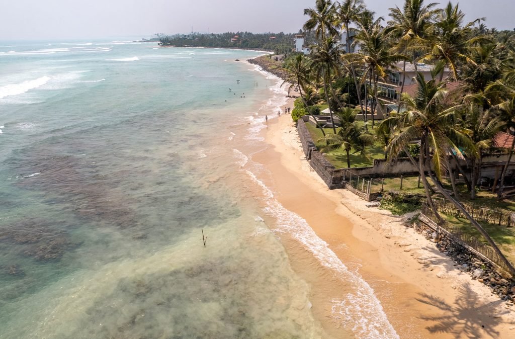 Aerial view of a coastal strip with a sandy beach edged by shallow coral reefs and lined with palm trees, adjacent to a coastal town. It is Kabalana Beach, one of the best beaches in Unawatuna.