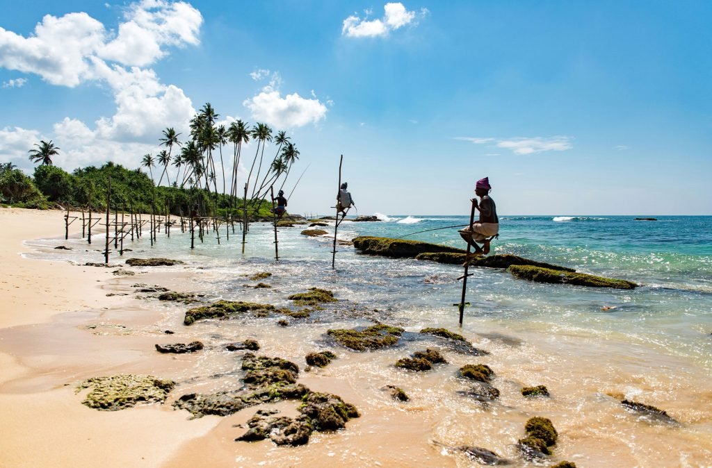 Traditional stilt fishermen perched on poles above shallow ocean waters near a tropical beach with palm trees in the background.
