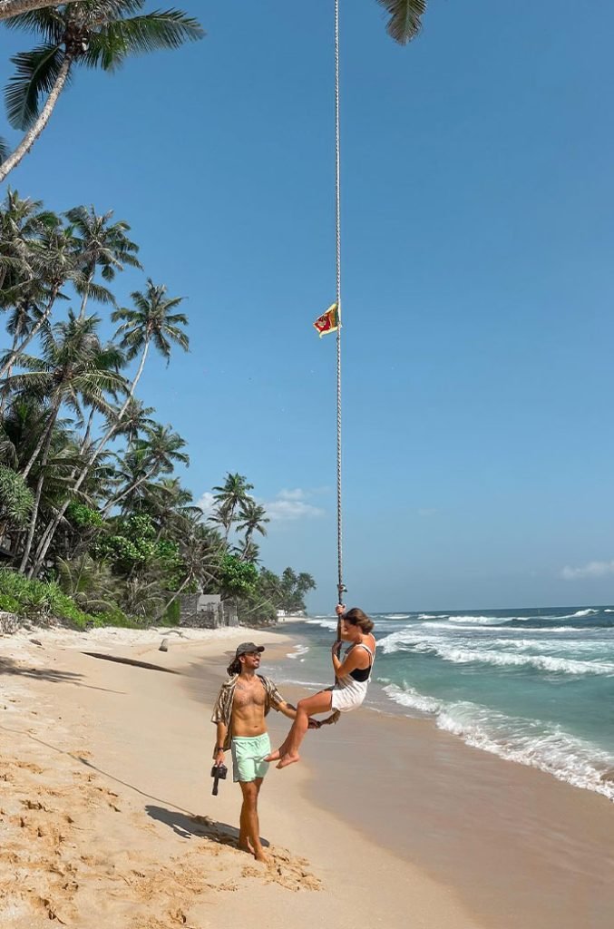 A person swinging on a beachside rope swing held by a local, with the Sri Lankan flag flying high above and the ocean's waves in the background