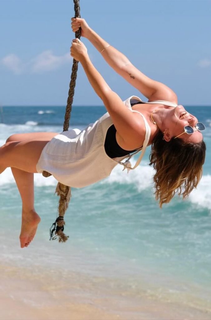 Joyful moment of a woman swinging on a beachside rope, mid-air with a carefree pose against the backdrop of turquoise waves