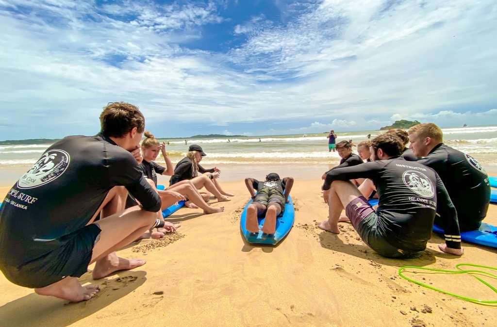A group of surfers in black rash guards sitting on surfboards on a sandy beach, attentively listening to a surfing lesson.