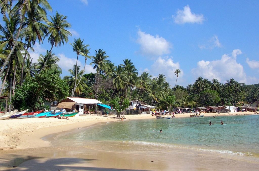A serene beach scene with a curve of golden sand lined by tropical palm trees and small boats, with a few people swimming in the clear, calm waters.
