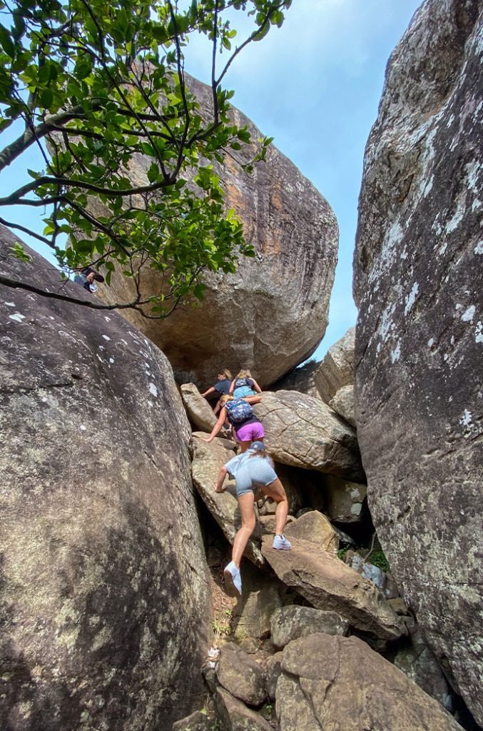 Adventurers climbing through a narrow passage between large boulders, surrounded by lush foliage