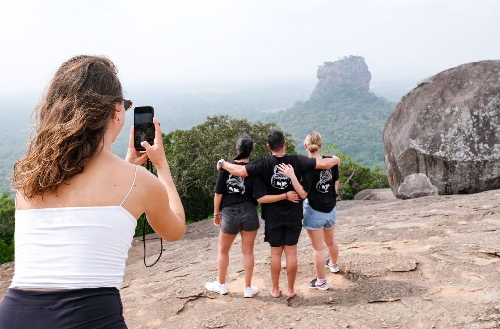 A woman captures a moment with her phone as a group admires a distant rock formation from a mountain viewpoint, embodying adventure and travel memories