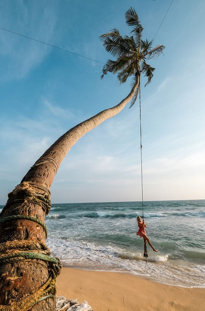 A person in a red dress swings from a rope tied to a palm tree leaning over a tropical beach with clear blue waters