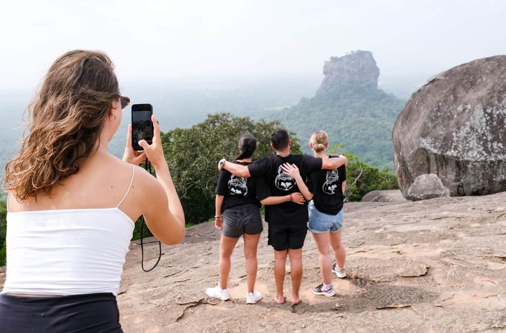 A woman in a white tank top using a smartphone to photograph three people wearing matching black t-shirts, looking out over a foggy mountain landscape. Is Sri Lanka Expensive? No.