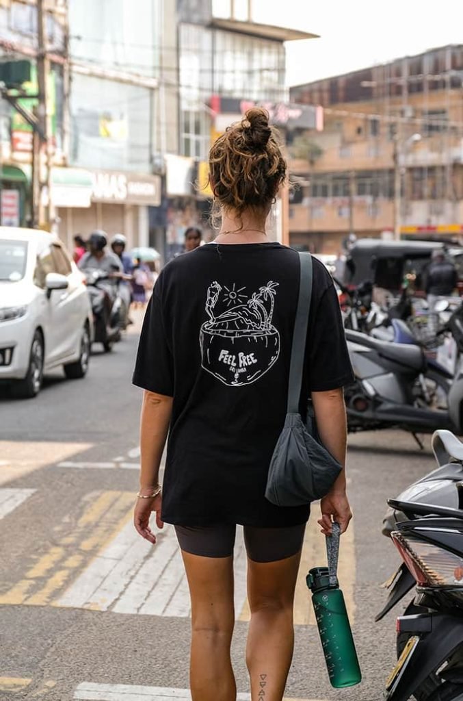 Rear view of a woman walking down a busy street; she is wearing a black t-shirt with a 'Feel Free Travel' logo and carrying a green water bottle.
