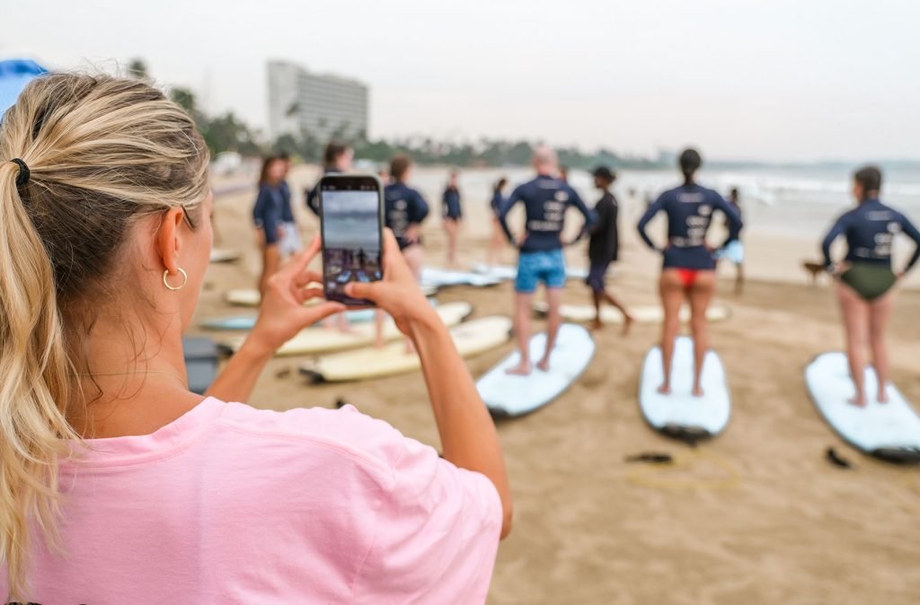 A woman in a pink t-shirt using her phone to capture a group of people preparing to surf, standing on the beach with surfboards