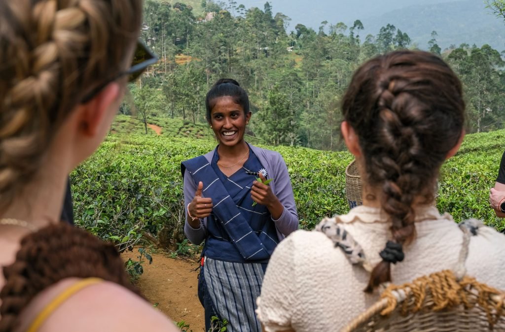 A smiling woman in traditional attire speaking with a group of tourists in a lush tea plantation; the tourists are listening attentively