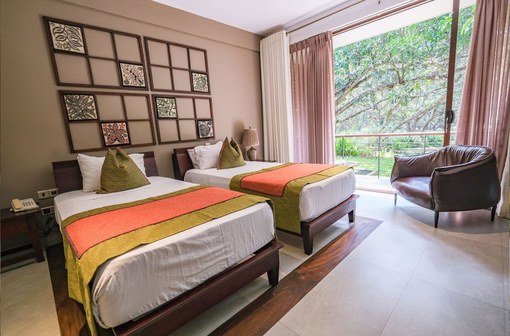 An elegant hotel room featuring twin beds with multicolored bedspreads, dark wood furnishings, and a large window overlooking lush greenery.
